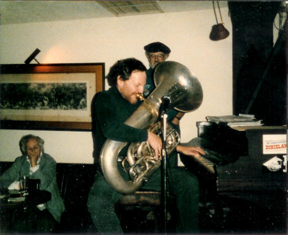Eli playing piano with left hand, tuba with right, Muriel Owen and Bob Pilsbury in background